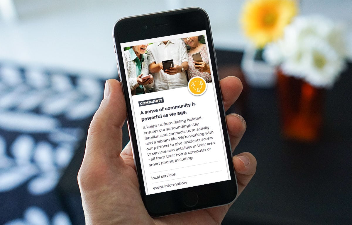 A hand holds a mobile phone displaying an article about community support for ageing, featuring a photo of people using mobile devices together.